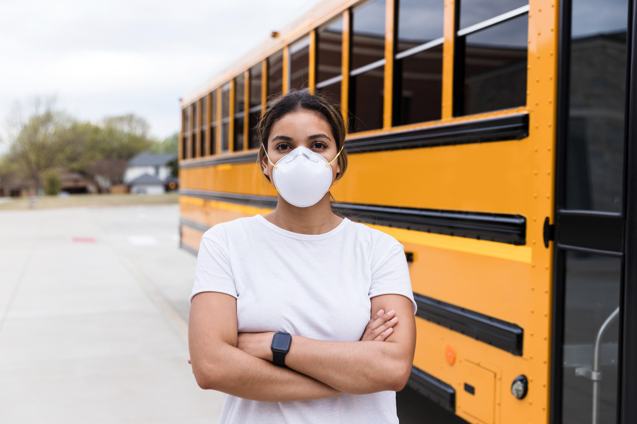 Person with face mask stands with arms crossed next to school bus.