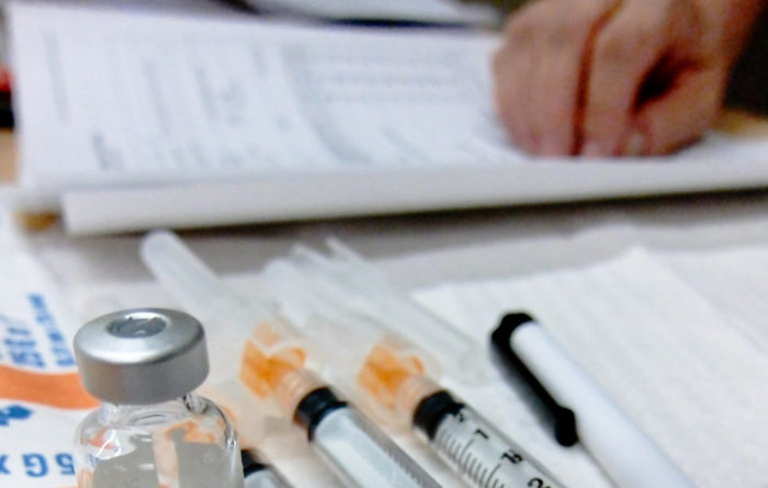 In the foreground are immunization plungers and vials. In the background is a hand resting on paper.