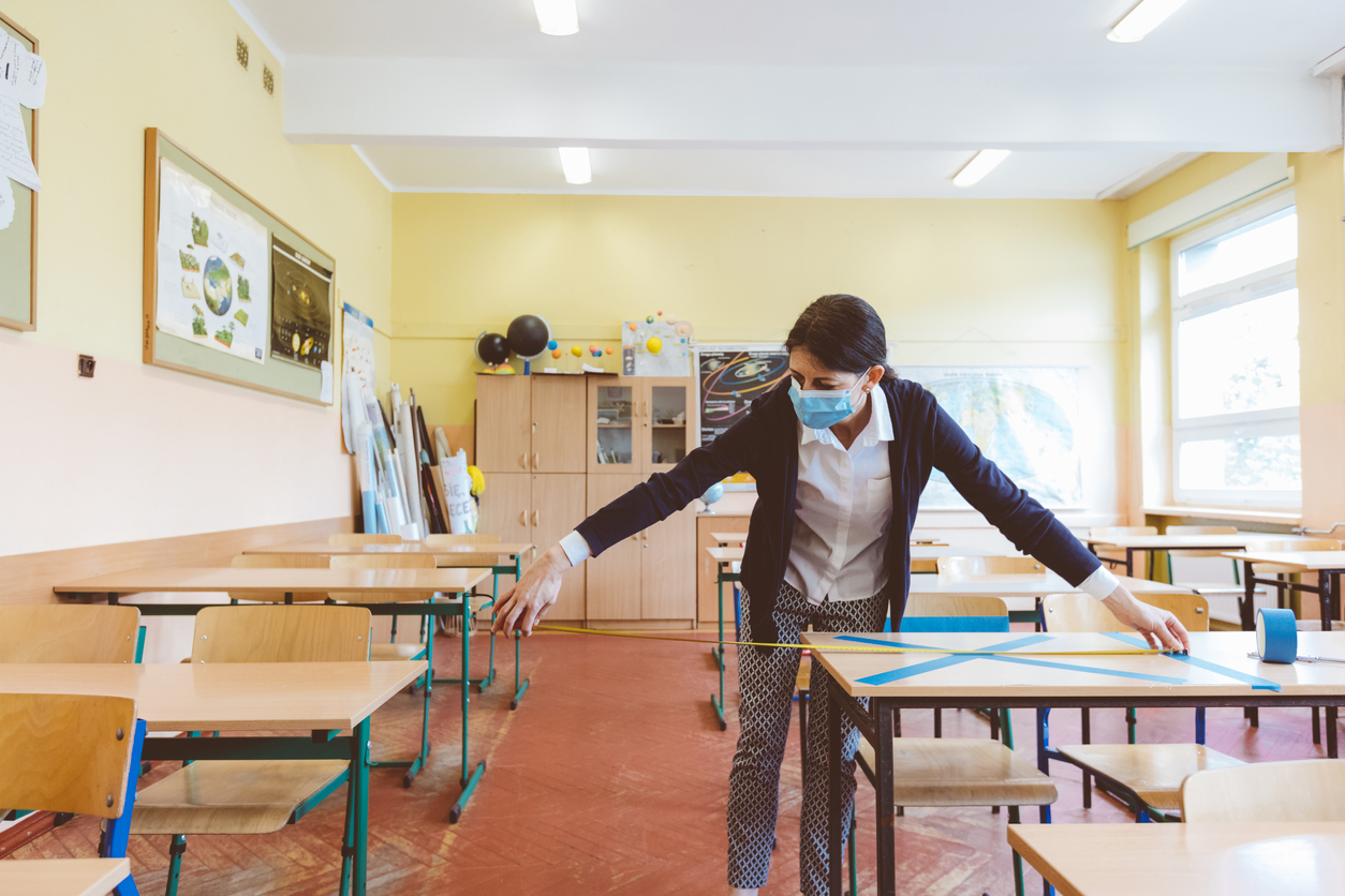 Teacher in a classroom, measuring distance between desks. The teacher is wearing a face mask.