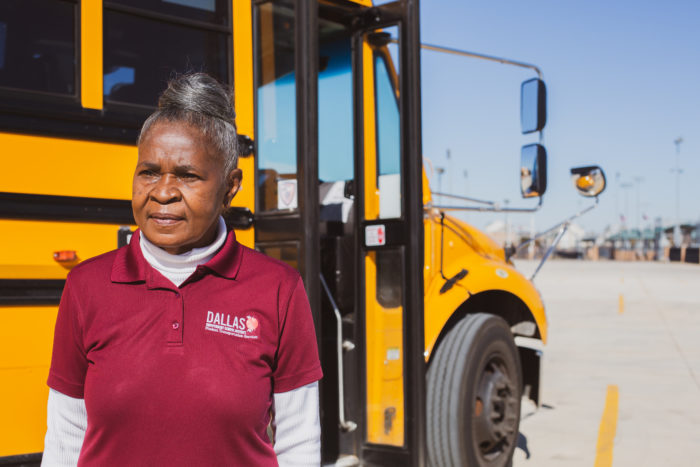 Member in Dallas I-S-D uniform standing by a school bus.