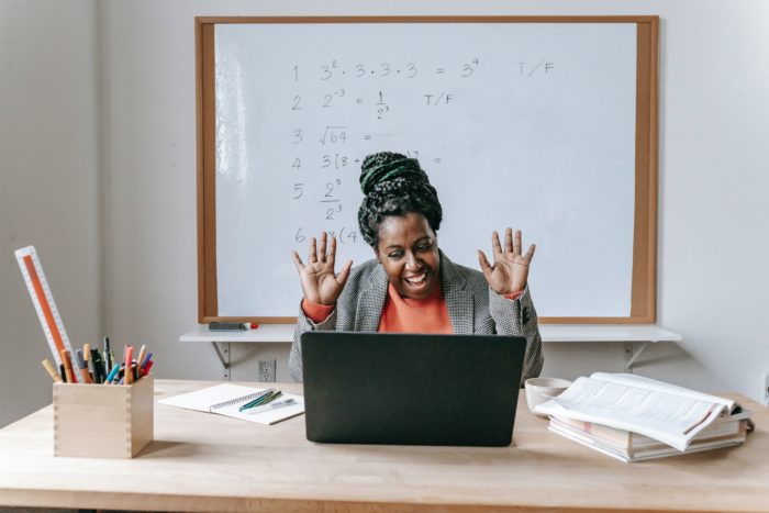 A teacher looks at their laptop while seated at a desk in a classroom. They are smiling and raising their hands.