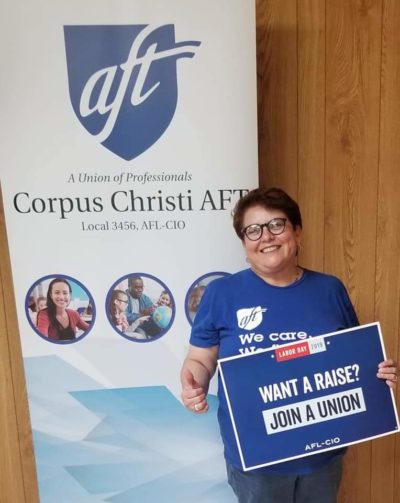 Doctor Nancy Vera stands in front of a Corpus Christi A-F-T banner. She holds a sign that reads, "Want a raise? Join a union."